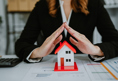 Hands forming a roof shape over a small model house on a desk with financial papers.